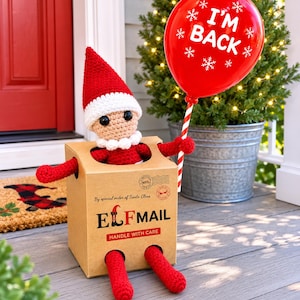 May include: A crocheted elf doll with a red and white hat sits inside a cardboard box labelled ELF MAIL. The box features the text By special order of Santa Claus and HANDLE WITH CARE. The elf holds a red balloon with white I'M BACK text.