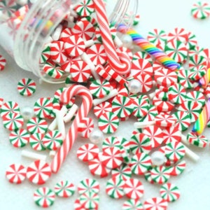 May include: A collection of Christmas-themed polymer clay pieces, including red and white peppermint candies, candy canes, and green and white striped candies. Scattered around are small white pearls and a rainbow-colored candy. The items are spilling from a clear jar.