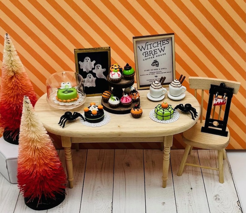 K&ouml;nnte beinhalten: Ein Miniatur-Halloween-Caf&eacute; mit einem Schild, auf dem "Witches Brew Coffee House" steht. Das Caf&eacute; hat einen Holztisch mit einer Auswahl an Miniaturkuchen und Cupcakes, die mit Halloween-Motiven verziert sind. Es gibt auch zwei Miniaturkaffeebecher und eine Miniaturguillotine.