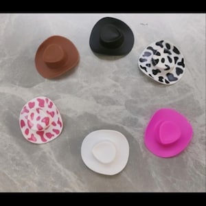 Puede incluir: Seis sombreros de vaquero en miniatura de varios colores y patrones, incluyendo negro, marrón, blanco con manchas negras, rosa y blanco con manchas rosas. Los sombreros están dispuestos en círculo sobre una superficie gris.