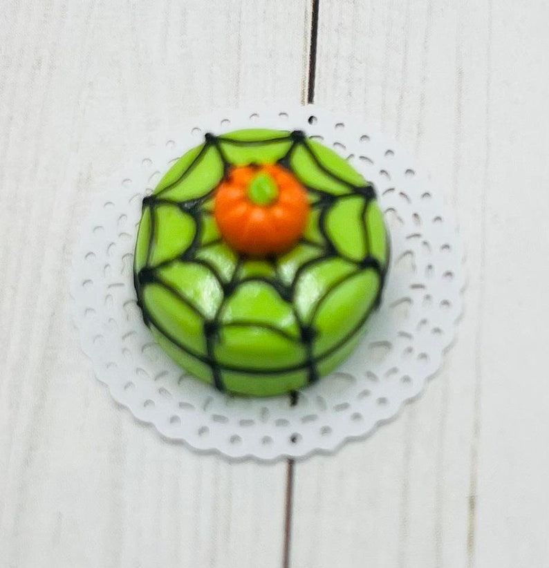 K&ouml;nnte beinhalten: Ein gr&uuml;ner und schwarzer Schokoladenkeks mit Spinnennetz-Muster und einem orangefarbenen K&uuml;rbis darauf.