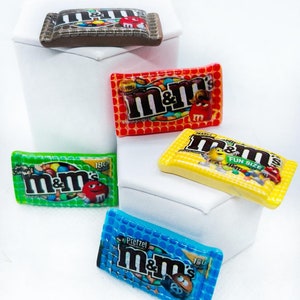May include: Four miniature M&M's candy bags in different colors and flavors: red, green, yellow, and blue. The bags are decorated with the M&M's logo and the flavor names.
