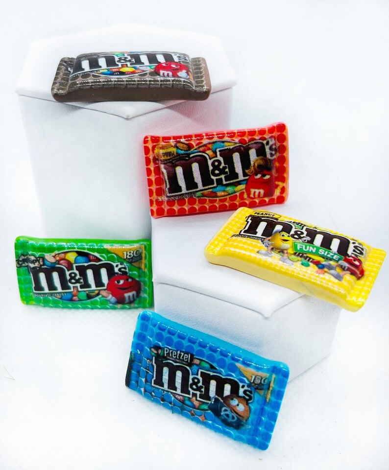 May include: Four miniature M&M's candy bags in different colors and flavors: red, green, yellow, and blue. The bags are decorated with the M&M's logo and the flavor names.