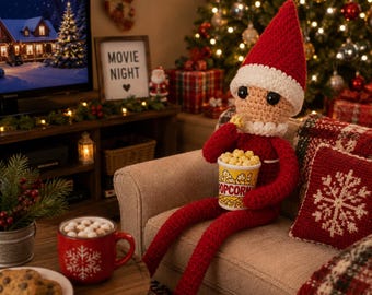 Könnte beinhalten: Eine gehäkelte Elfenpuppe mit roter Mütze und Anzug, weißem Besatz und schwarzen Augen, die auf einem Sofa sitzt und einen kleinen weißen Eimer mit der Aufschrift POPCORN und gelbem Popcorn hält, wobei ein Stück nahe am Mund ist.
