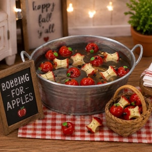 May include: A miniature galvanized metal tub filled with water, containing several red apples and apple cores. A small wooden sign reads BOBBING FOR APPLES with a red apple graphic. A woven basket holds more apples and cores on a red and white checkered cloth.
