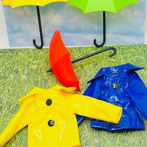 May include: A set of three miniature umbrellas in yellow, green, and red, along with two miniature raincoats in yellow and blue. The umbrellas and raincoats are displayed on a green grassy surface.