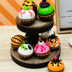 K&ouml;nnte beinhalten: Ein zweist&ouml;ckiger Kuchenst&auml;nder aus Holz mit einer Auswahl an Halloween-Cupcakes. Die Cupcakes sind mit orangefarbenem, gr&uuml;nem, rosa, wei&szlig;em und schwarzem Frosting verziert und haben verschiedene Halloween-Designs, darunter Spinnen, K&uuml;rbisse und Geister.