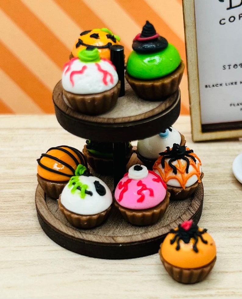 K&ouml;nnte beinhalten: Ein zweist&ouml;ckiger Kuchenst&auml;nder aus Holz mit einer Auswahl an Halloween-Cupcakes. Die Cupcakes sind mit orangefarbenem, gr&uuml;nem, rosa, wei&szlig;em und schwarzem Frosting verziert und haben verschiedene Halloween-Designs, darunter Spinnen, K&uuml;rbisse und Geister.