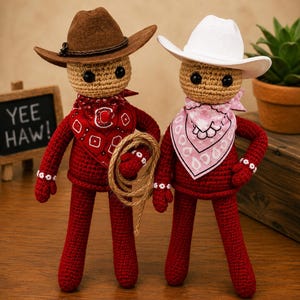 Puede incluir: Dos muñecos de ganchillo con cabezas color canela y cuerpos rojos. Uno lleva un sombrero de vaquero marrón y un pañuelo rojo con una letra C, sosteniendo una cuerda. El otro lleva un sombrero de vaquero blanco y un pañuelo rosa estampado.