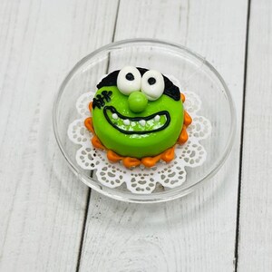 K&ouml;nnte beinhalten: Ein gr&uuml;ner und wei&szlig;er Kuchen, der mit einem Monstergesicht, schwarzen Augen und einem wei&szlig;en L&auml;cheln verziert ist. Der Kuchen befindet sich auf einer wei&szlig;en Spitze und einem durchsichtigen Glasteller.