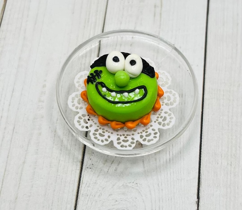 K&ouml;nnte beinhalten: Ein gr&uuml;ner und wei&szlig;er Kuchen, der mit einem Monstergesicht, schwarzen Augen und einem wei&szlig;en L&auml;cheln verziert ist. Der Kuchen befindet sich auf einer wei&szlig;en Spitze und einem durchsichtigen Glasteller.