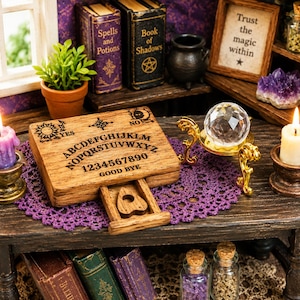 Op de afbeelding: Een houten spirit board met een schuiflade met een planchette, geplaatst op een paars kanten kleedje. Het bord toont het alfabet, de cijfers nul tot en met negen, de woorden YES, NO, GOOD BYE en een kristallen bol op een gouden standaard.