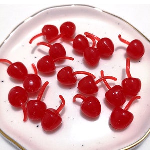 May include: A selection of small, shiny red cherry-shaped beads with curved red stems, arranged on a white plate with a gold rim. The beads are evenly spaced, creating a pleasing display. The cherries are a bright red colour.