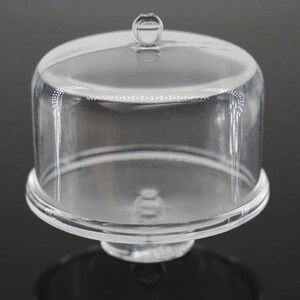 K&ouml;nnte beinhalten: Eine transparente Kuchenhaube aus Kunststoff mit einem runden Sockel und einem Knopf oben. Die Haube ist dazu bestimmt, Kuchen oder andere Desserts abzudecken und zu sch&uuml;tzen.