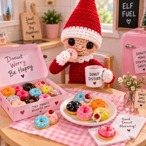 May include: A crocheted elf doll with a red hat and white trim holds a miniature doughnut and a white mug labelled Donut Disturb. A pink box labelled Donut Worry Be Happy contains assorted miniature doughnuts, with more doughnuts on a white plate.