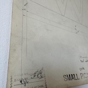 Pode incluir: Um desenho técnico vintage em papel branco sujo com o contorno de uma letra W grande, anotações manuscritas, uma secção carimbada com MADE BY e EXAM BY com assinaturas, além do texto impresso SMALL PICA e Head of Dept.