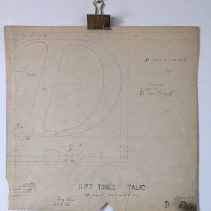 Pode incluir: Um esboço de planta vintage da letra "D" na fonte Times Italic, preso por um clipe de latão. O papel envelhecido apresenta notas manuscritas, medições e o texto "II PT TIMES ITALIC". O esboço é montado em uma parede branca.