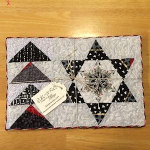 May include: A quilted table runner featuring a star design in black, white and grey fabric. The runner is tied with twine and has a tag that reads "Quilt-a-Rie! Designs by Marie Jaeger QuiltanieEtsy.etsy.com MarieCreativeSpace.blogspot.com"