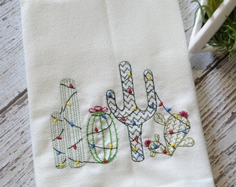 Puede incluir: Toalla de cocina blanca con un diseño bordado de tres cactus con luces navideñas coloridas envueltas alrededor.
