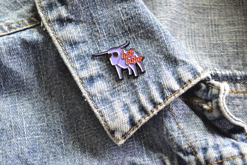 Puede incluir: Pin de esmalte morado de un toro con el texto "hot babe" en letras rojas. El pin est&aacute; en una chaqueta vaquera azul.