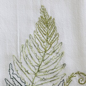 May include: Embroidered fern leaf design on a white fabric kitchen towel. The fern is outlined in black thread and filled in with green thread.