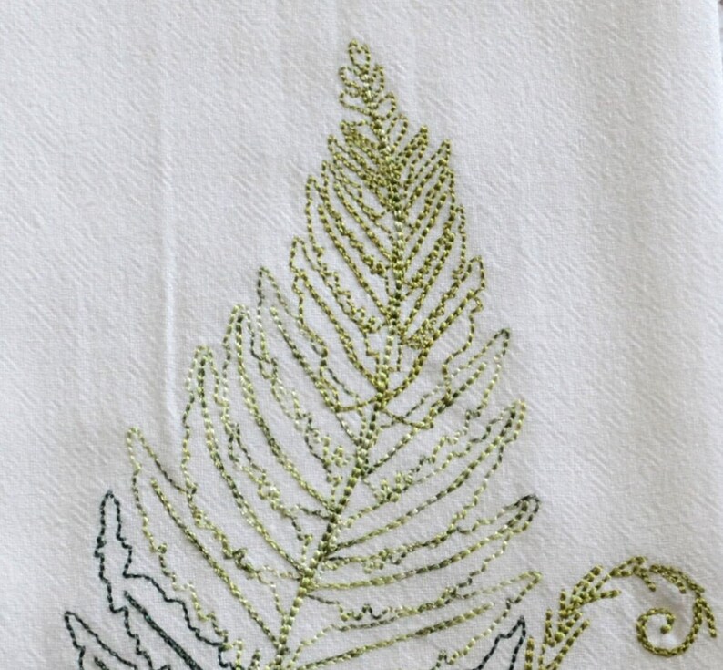 May include: Embroidered fern leaf design on a white fabric kitchen towel. The fern is outlined in black thread and filled in with green thread.