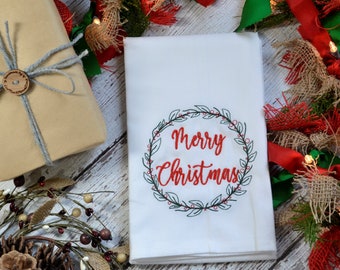 Puede incluir: Toalla de cocina blanca con el texto bordado en rojo "Merry Christmas" dentro de una corona verde con bayas rojas.