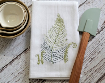 May include: White kitchen towel with an embroidered green fern design. A green silicone spatula with a wooden handle is lying on top of the towel.