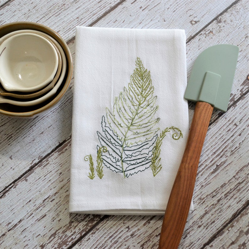 May include: White kitchen towel with an embroidered green fern design. A green silicone spatula with a wooden handle is lying on top of the towel.
