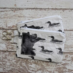 May include: Two zippered pouches with a white background and black loon print. The pouches are sewn together and have a black outline of the state of Minnesota on the front.