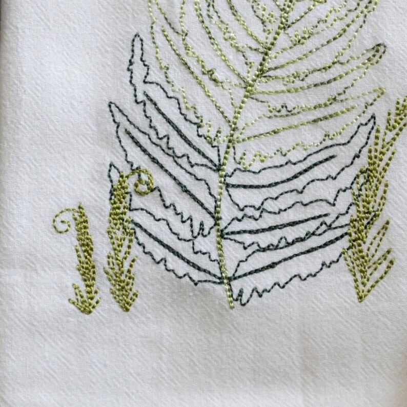 May include: Embroidered green and black fern leaves on a white fabric background.
