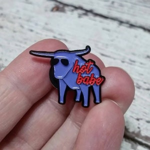 Puede incluir: Pin de esmalte morado de un toro con gafas de sol con el texto "hot babe" en rojo.
