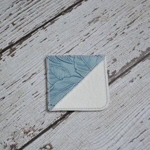 May include: A blue and white fabric bookmark with a floral pattern. The bookmark is folded in half, with the blue side facing up and the white side facing down.