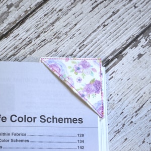 May include: A pink and purple floral fabric bookmark with a white background. The bookmark is in the shape of a triangle and has pink stitching around the edges.