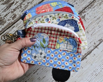 May include: A collection of handmade fabric coin purses in various floral and patterned designs. The purses feature a camper design with a zipper closure and a key ring attachment. The color palette includes blue, red, and white.