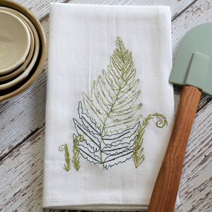 May include: White kitchen towel with an embroidered green fern design. The towel is folded in half and has a green and brown spatula next to it.