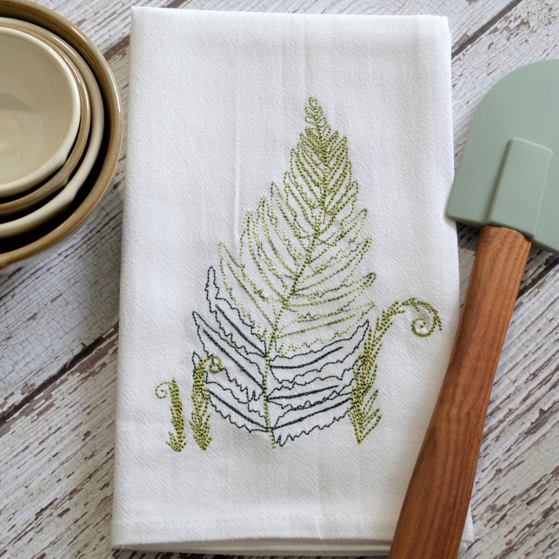 May include: White kitchen towel with an embroidered green fern design. The towel is folded in half and has a green and brown spatula next to it.