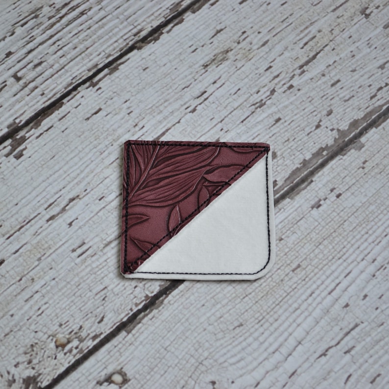 May include: A burgundy leather bookmark with a floral pattern on one side and a white fabric on the other side.