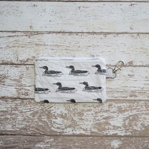 May include: A white zippered pouch with a quilted design and a black and white loon pattern. The pouch has a silver zipper pull and a silver key ring.