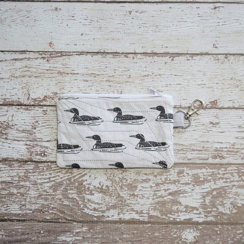 May include: A white zippered pouch with a quilted design and a black and white loon pattern. The pouch has a silver zipper pull and a silver key ring.