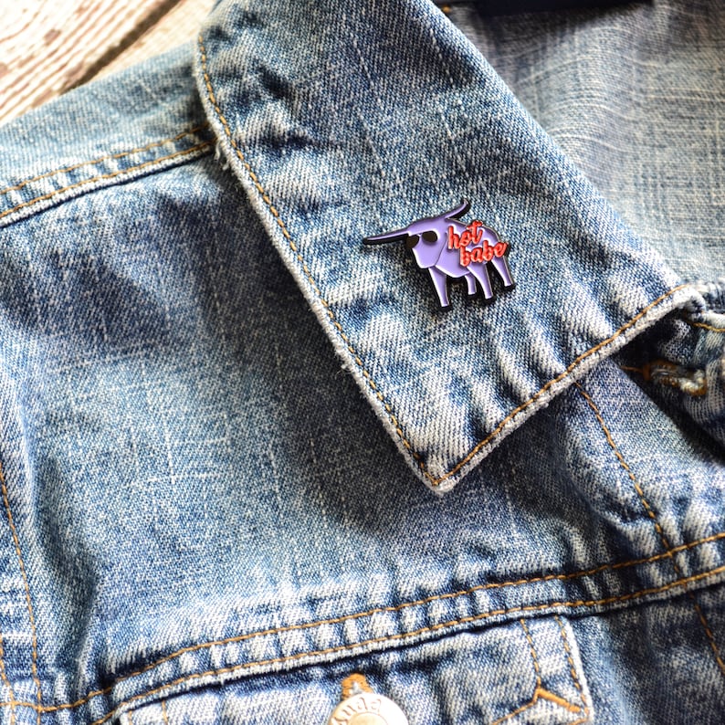 Puede incluir: Un pin de esmalte morado con un toro de dibujos animados y el texto "hot babe" en letras rojas. El pin est&aacute; sujeto a una chaqueta vaquera azul.