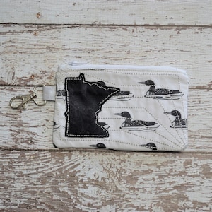 May include: A white zippered pouch with a black outline of the state of Minnesota and a pattern of black loon birds on a white background.