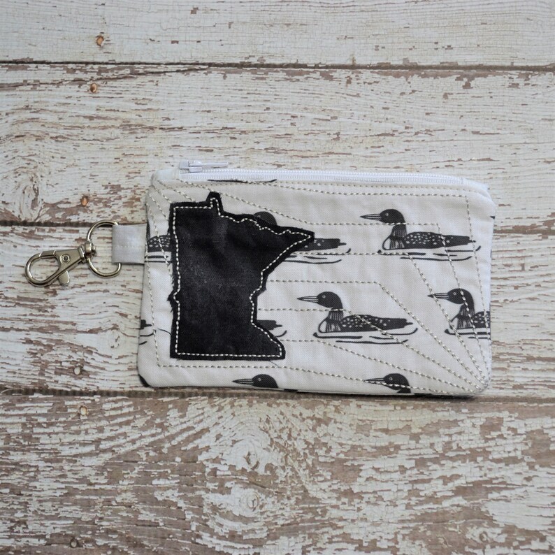 May include: A white zippered pouch with a black outline of the state of Minnesota and a pattern of black loon birds on a white background.
