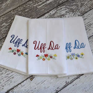May include: Three white kitchen towels with embroidered floral designs and the phrase "Uff Da" in different colors. The towels are folded and arranged on a wooden surface.