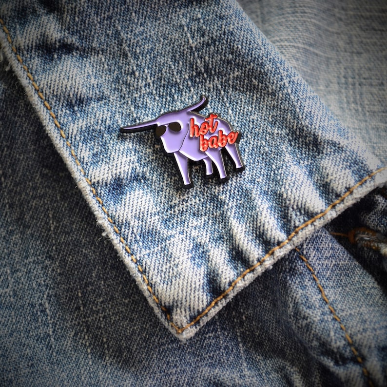 Puede incluir: Un pin de esmalte morado de un toro con el texto "hot babe" en letras rojas.
