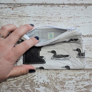 May include: A white zippered pouch with a black and white loon pattern. The pouch is open and the inside is lined with white fabric. The zipper pull has a small white tag with the text "Sand Black More" on it.