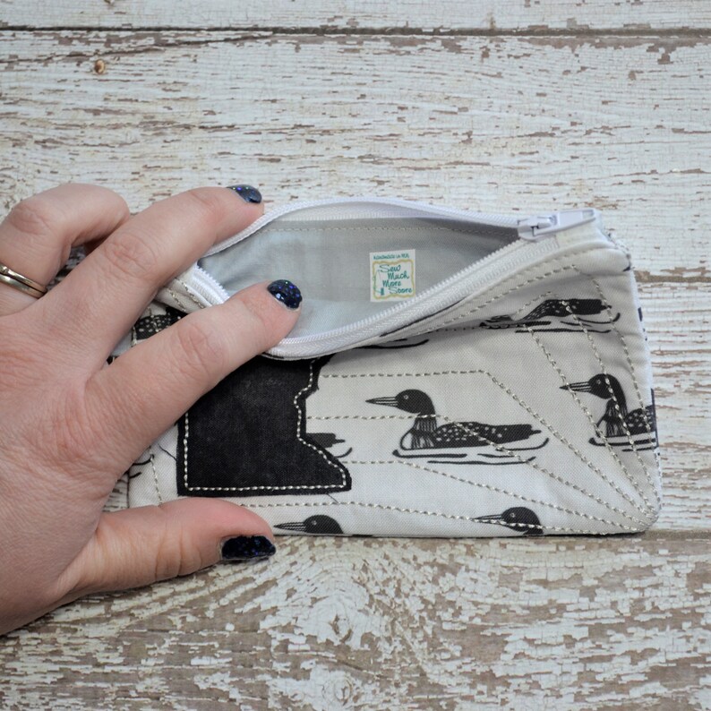 May include: A white zippered pouch with a black and white loon pattern. The pouch is open and the inside is lined with white fabric. The zipper pull has a small white tag with the text "Sand Black More" on it.