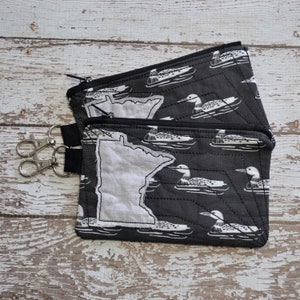 May include: Two black zippered pouches with a white outline of the state of Minnesota and a pattern of white loon birds on a black background.