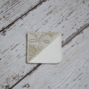 May include: A beige leather bookmark with a floral pattern on one side and a plain beige fabric on the other side.