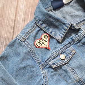 May include: A blue denim jacket with a red heart-shaped patch that says "Uffda" in white embroidery.